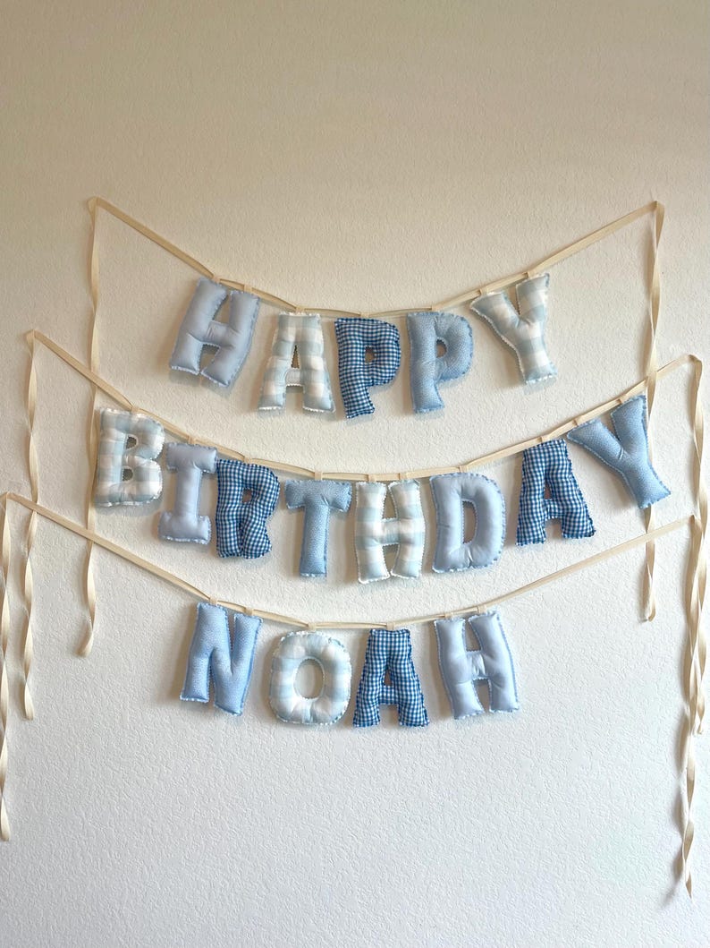 K&ouml;nnte beinhalten: Ein dreistufiges Stoffbanner mit ausgestopften, weichen Buchstaben in verschiedenen Blau- und Wei&szlig;t&ouml;nen. Die Buchstaben bilden den Schriftzug "HAPPY BIRTHDAY NOAH" und sind an d&uuml;nnen, cremefarbenen B&auml;ndern befestigt. Jeder Buchstabe hat ein Muster.