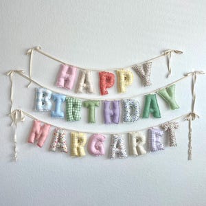K&ouml;nnte beinhalten: Ein dreireihiges Stoffbanner mit ausgestopften, einzelnen Buchstaben, die HAPPY BIRTHDAY MARGARET bilden. Jeder Buchstabe ist aus verschiedenen pastellfarbenen Stoffen gefertigt, darunter Vichy-Karos, Blumenmuster und unifarbene Stoffe, und an d&uuml;nnen cremefarbenen B&auml;ndern befestigt.