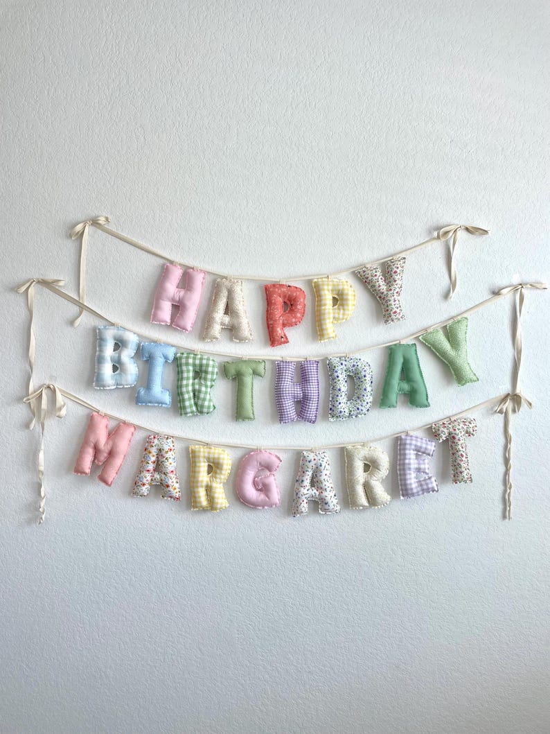K&ouml;nnte beinhalten: Ein dreireihiges Stoffbanner mit ausgestopften, einzelnen Buchstaben, die HAPPY BIRTHDAY MARGARET bilden. Jeder Buchstabe ist aus verschiedenen pastellfarbenen Stoffen gefertigt, darunter Vichy-Karos, Blumenmuster und unifarbene Stoffe, und an d&uuml;nnen cremefarbenen B&auml;ndern befestigt.