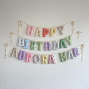 K&ouml;nnte beinhalten: Ein dreistufiges Stoffbuchstaben-Banner mit der Aufschrift HAPPY BIRTHDAY AURORA MAE. Jeder Buchstabe ist einzeln gef&uuml;llt und weist verschiedene pastellfarbene Muster auf, darunter Vichy-Karos, Blumen und einfarbige Stoffe, befestigt an cremefarbenen B&auml;ndern.