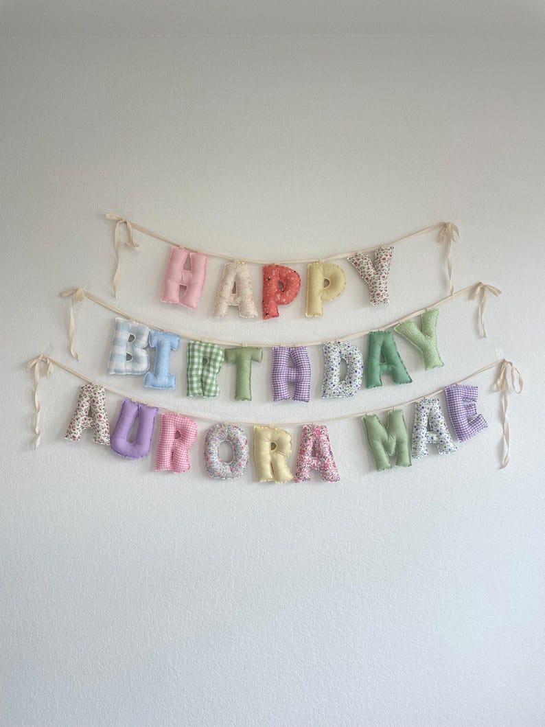 K&ouml;nnte beinhalten: Ein dreistufiges Stoffbuchstaben-Banner mit der Aufschrift HAPPY BIRTHDAY AURORA MAE. Jeder Buchstabe ist einzeln gef&uuml;llt und weist verschiedene pastellfarbene Muster auf, darunter Vichy-Karos, Blumen und einfarbige Stoffe, befestigt an cremefarbenen B&auml;ndern.
