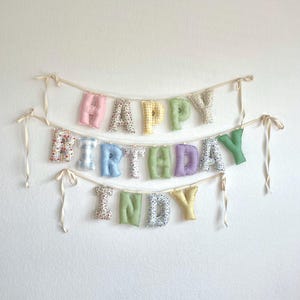 K&ouml;nnte beinhalten: Ein Stoffbanner mit drei Reihen aus ausgestopften, weichen Buchstaben, die HAPPY BIRTHDAY INDY buchstabieren. Die Buchstaben sind aus verschiedenen pastellfarbenen Stoffen mit Vichy-Karo-, Blumen- und einfarbigen Mustern gefertigt und an d&uuml;nnen cremefarbenen B&auml;ndern befestigt.