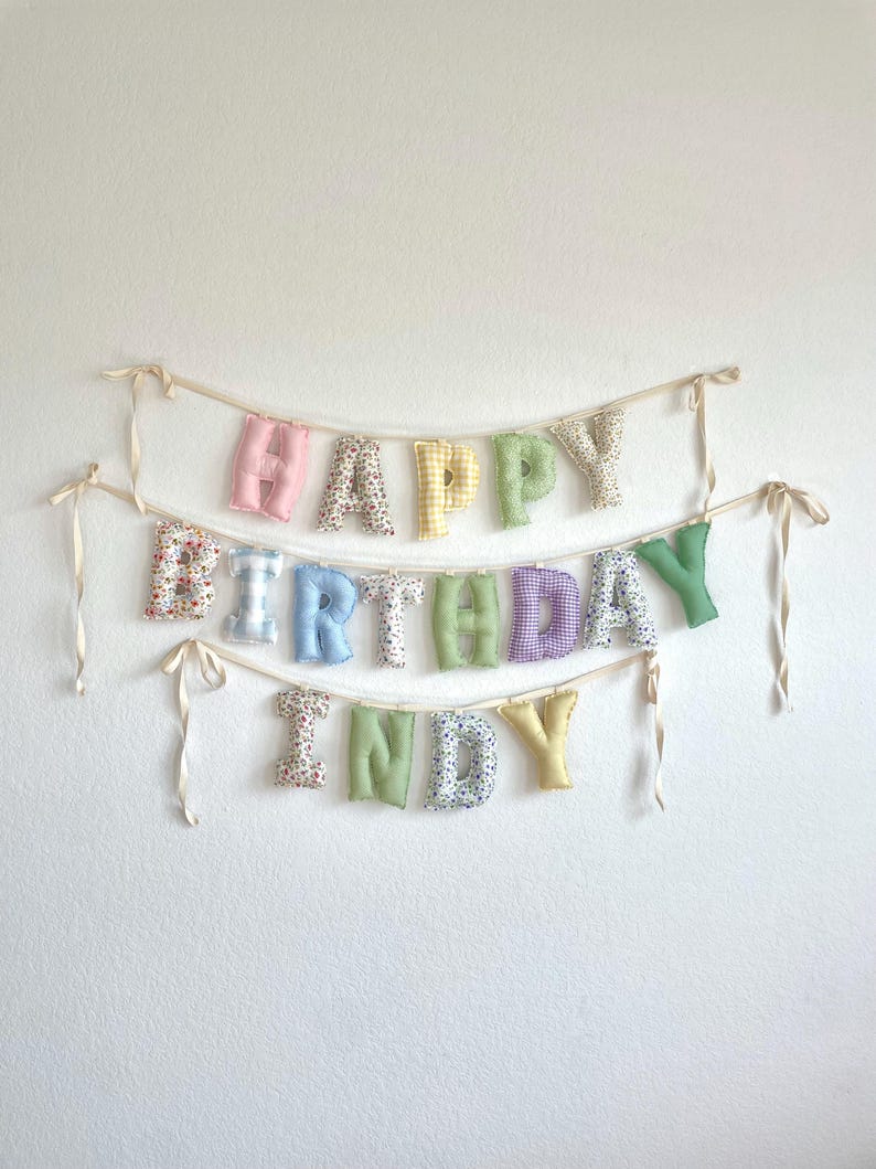 K&ouml;nnte beinhalten: Ein Stoffbanner mit drei Reihen aus ausgestopften, weichen Buchstaben, die HAPPY BIRTHDAY INDY buchstabieren. Die Buchstaben sind aus verschiedenen pastellfarbenen Stoffen mit Vichy-Karo-, Blumen- und einfarbigen Mustern gefertigt und an d&uuml;nnen cremefarbenen B&auml;ndern befestigt.