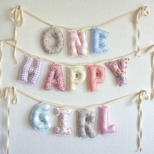 K&ouml;nnte beinhalten: Ein dekoratives Banner mit ausgestopften Stoffbuchstaben, die die Worte ONE HAPPY GIRL bilden. Die Buchstaben sind an cremefarbenen B&auml;ndern befestigt und zeigen verschiedene Muster, darunter Blumenprints, rosa-wei&szlig;es Vichy-Karo und Pastellt&ouml;ne.