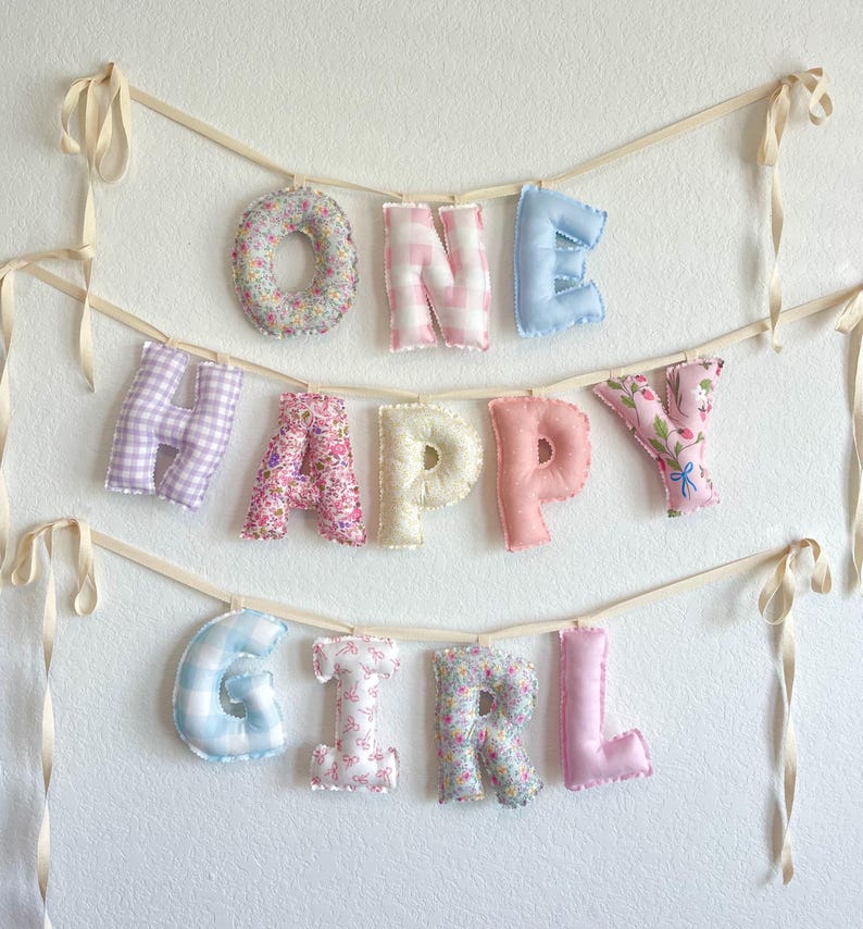 K&ouml;nnte beinhalten: Ein dekoratives Banner mit ausgestopften Stoffbuchstaben, die die Worte ONE HAPPY GIRL bilden. Die Buchstaben sind an cremefarbenen B&auml;ndern befestigt und zeigen verschiedene Muster, darunter Blumenprints, rosa-wei&szlig;es Vichy-Karo und Pastellt&ouml;ne.