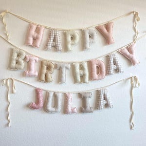 K&ouml;nnte beinhalten: Ein zweistufiges Stoffbanner mit ausgestopften, weichen Buchstaben, die "HAPPY BIRTHDAY JULIA" bilden. Die Buchstaben sind in verschiedenen Mustern gestaltet, darunter einfarbiges Rosa, Beige und Wei&szlig; sowie Vichy-Karos und florale Drucke, befestigt an cremefarbenen B&auml;ndern.