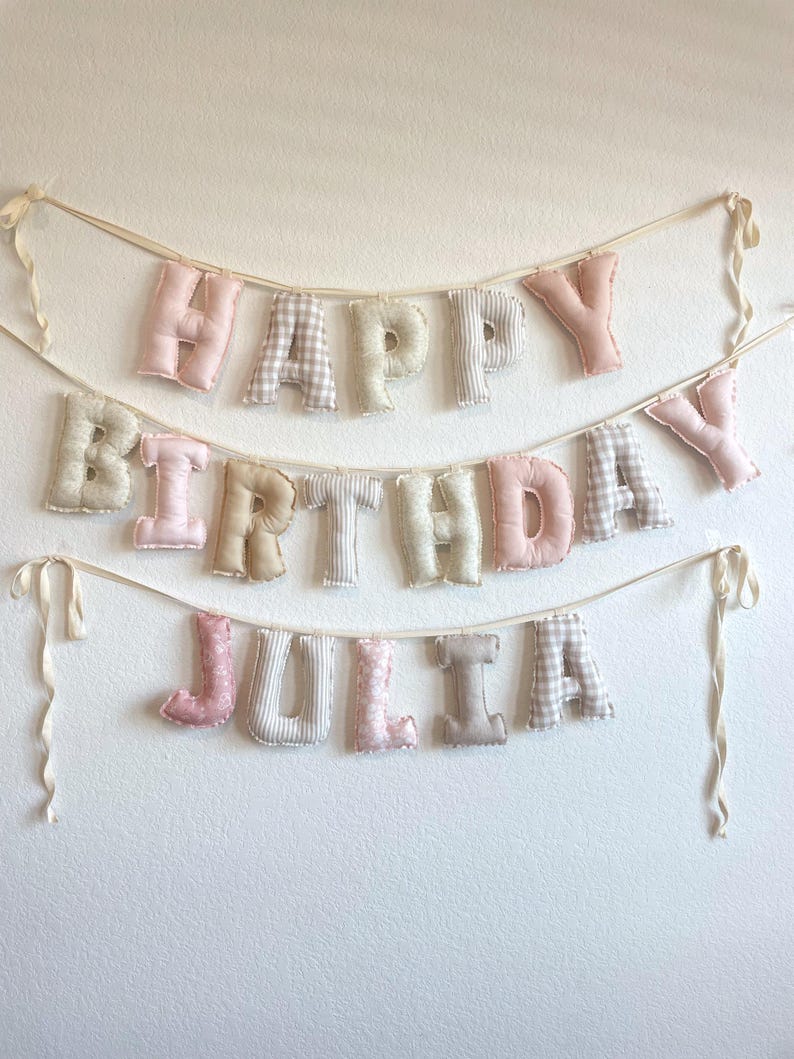 K&ouml;nnte beinhalten: Ein zweistufiges Stoffbanner mit ausgestopften, weichen Buchstaben, die "HAPPY BIRTHDAY JULIA" bilden. Die Buchstaben sind in verschiedenen Mustern gestaltet, darunter einfarbiges Rosa, Beige und Wei&szlig; sowie Vichy-Karos und florale Drucke, befestigt an cremefarbenen B&auml;ndern.
