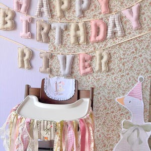 K&ouml;nnte beinhalten: Ein Stoffbanner mit der Aufschrift HAPPY BIRTHDAY RIVER aus gepolsterten Pastellbuchstaben, ein wei&szlig;es L&auml;tzchen mit R&uuml;schen, der Zahl 1 und dem Namen River sowie ein Hochstuhl-Rock aus rosa, wei&szlig;en und floralen B&auml;ndern mit G&auml;nsemotiv.
