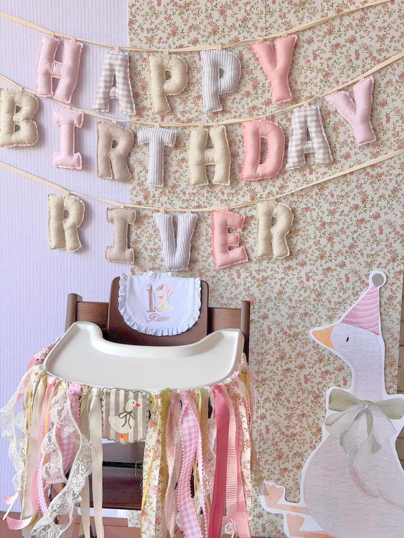 K&ouml;nnte beinhalten: Ein Stoffbanner mit der Aufschrift HAPPY BIRTHDAY RIVER aus gepolsterten Pastellbuchstaben, ein wei&szlig;es L&auml;tzchen mit R&uuml;schen, der Zahl 1 und dem Namen River sowie ein Hochstuhl-Rock aus rosa, wei&szlig;en und floralen B&auml;ndern mit G&auml;nsemotiv.