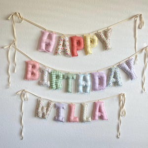 K&ouml;nnte beinhalten: Ein dreistufiges Stoffbanner mit ausgestopften, weichen Buchstaben, die HAPPY BIRTHDAY WILLA buchstabieren. Die Buchstaben bestehen aus verschiedenen pastellfarbenen Stoffen mit Blumen-, Vichy- und einfarbigen Mustern und h&auml;ngen an schmalen cremefarbenen B&auml;ndern.