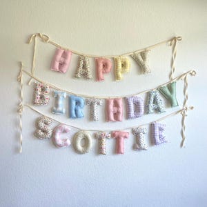 K&ouml;nnte beinhalten: Ein dreistufiges Stoffbanner mit der Aufschrift HAPPY BIRTHDAY SCOTTIE. Jeder Buchstabe ist ein ausgestopfter, einzelner Pl&uuml;schcharakter mit verschiedenen Pastellfarben und Mustern, darunter Blumen, Vichy-Karo sowie einfarbiges Rosa, Blau, Gelb und Gr&uuml;n.