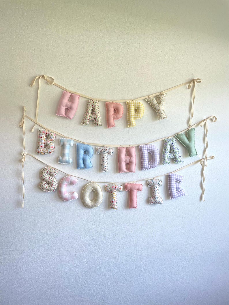K&ouml;nnte beinhalten: Ein dreistufiges Stoffbanner mit der Aufschrift HAPPY BIRTHDAY SCOTTIE. Jeder Buchstabe ist ein ausgestopfter, einzelner Pl&uuml;schcharakter mit verschiedenen Pastellfarben und Mustern, darunter Blumen, Vichy-Karo sowie einfarbiges Rosa, Blau, Gelb und Gr&uuml;n.