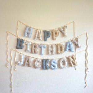 K&ouml;nnte beinhalten: Ein dreistufiges Stoffbanner mit ausgestopften, weichen Buchstaben, die "HAPPY BIRTHDAY JACKSON" bilden. Die Buchstaben sind in verschiedenen Mustern und Farben wie Hellblau, Beige und Grau gehalten und an d&uuml;nnen, cremefarbenen B&auml;ndern befestigt.