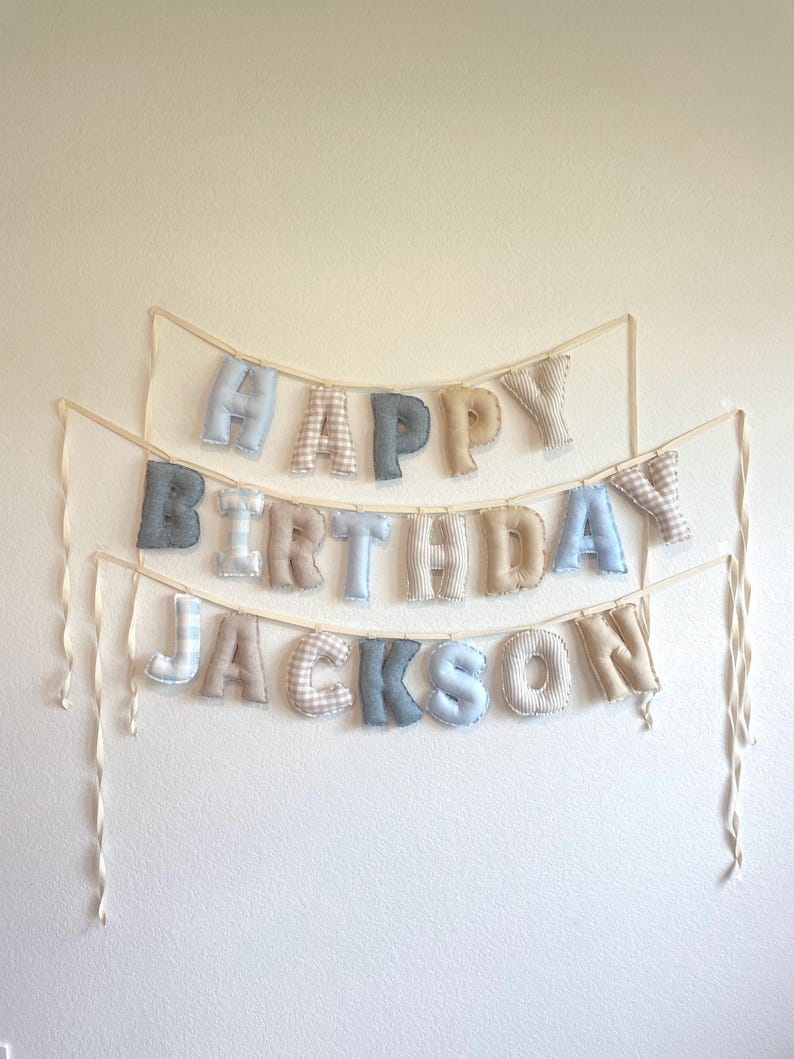 K&ouml;nnte beinhalten: Ein dreistufiges Stoffbanner mit ausgestopften, weichen Buchstaben, die "HAPPY BIRTHDAY JACKSON" bilden. Die Buchstaben sind in verschiedenen Mustern und Farben wie Hellblau, Beige und Grau gehalten und an d&uuml;nnen, cremefarbenen B&auml;ndern befestigt.