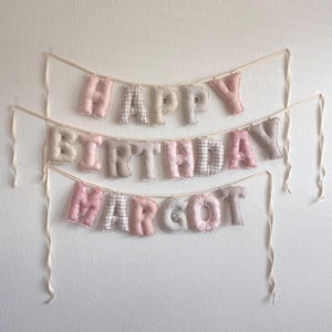 K&ouml;nnte beinhalten: Ein dreireihiges Stoffbanner mit der Aufschrift HAPPY BIRTHDAY MARGOT. Die ausgestopften, weichen Buchstaben bestehen aus einer Mischung aus einfarbig rosa, beigen und grauen Stoffen sowie Karomustern, Streifen und Punkten, befestigt an cremefarbenen B&auml;ndern.