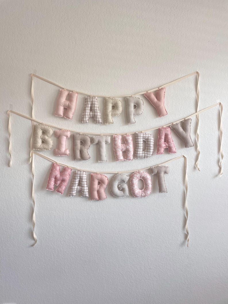 K&ouml;nnte beinhalten: Ein dreireihiges Stoffbanner mit der Aufschrift HAPPY BIRTHDAY MARGOT. Die ausgestopften, weichen Buchstaben bestehen aus einer Mischung aus einfarbig rosa, beigen und grauen Stoffen sowie Karomustern, Streifen und Punkten, befestigt an cremefarbenen B&auml;ndern.