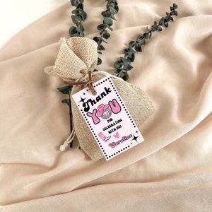 Puede incluir: Una pequeña bolsa de regalo de arpillera atada con una cuerda marrón, con una etiqueta rosa que dice "Thank You for Celebrating with Me!" en una fuente retro. La etiqueta presenta un sombrero de vaquero, una bola de discoteca y un gráfico de bota de vaquero. Ramas de eucalipto en el fondo.