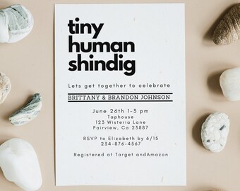 May include: A white invitation card with the text "tiny human shindig" in bold black letters, surrounded by various smooth stones on a beige background. The invitation includes event details.