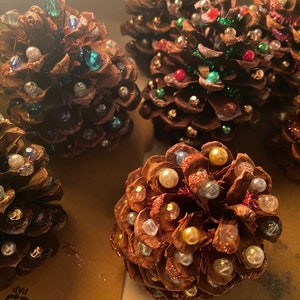 May include: Close-up of several decorated pine cones. The pine cones are brown and adorned with various colorful beads and pearls. Some cones have green tinsel accents. The decorations give the pine cones a festive appearance.