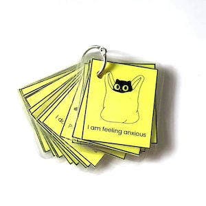 Puede incluir: Un conjunto de tarjetas cuadradas amarillas unidas por un anillo de metal plateado. Cada tarjeta presenta un dibujo lineal negro de un gato dentro de una bolsa y el texto I am feeling anxious. Las tarjetas están plastificadas y en abanico.