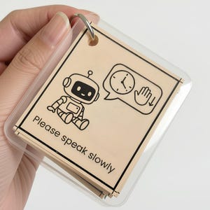 May include: A square communication card with a light beige background and a black border. It features a cartoon robot, a speech bubble containing a clock and a hand icon, and the printed text "Please speak slowly" in a clear black font.