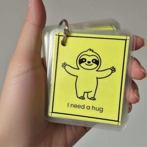 May include: A stack of square yellow cards held together by a silver metal ring. Each card features a black line drawing of a sloth with its arms outstretched and the printed text "I need a hug" at the bottom, all encased in clear plastic.