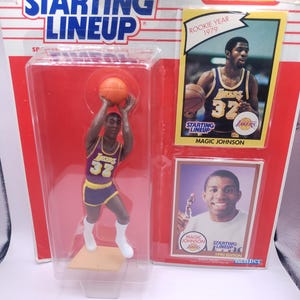 Puede incluir: Una figura de Starting Lineup de Magic Johnson con uniforme de los Lakers, sosteniendo un balón de baloncesto. El embalaje incluye una tarjeta Rookie Year 1979 y una tarjeta de la edición de 1990. La figura está en una caja de plástico transparente con fondo rojo.