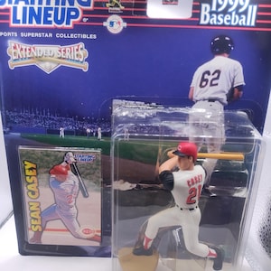 Puede incluir: Una figura de béisbol Starting Lineup de 1999 de Sean Casey con el uniforme de los Rojos de Cincinnati, empaquetada con una tarjeta coleccionable. La figura está en pose de bateo, con gorra roja y el número 21 en la camiseta. El embalaje dice "Extended Series".