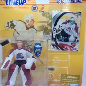 Może przedstawiać: Figurka kolekcjonerska Starting Lineup z 1998 roku przedstawiająca Patricka Roya, bramkarza hokejowego, w jego stroju Colorado Avalanche. Figurka znajduje się w przezroczystym plastikowym opakowaniu z kartą kolekcjonerską. Opakowanie zawiera tekst "Starting Lineup" i "1998 Edition".