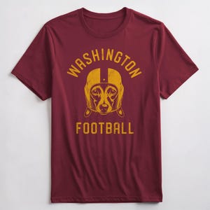 May include: Burgundy t-shirt with a yellow and gold graphic of a dog wearing a football helmet. The text "WASHINGTON FOOTBALL" is printed on the shirt.