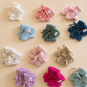 May include: A collection of twelve pairs of knitted baby booties in various colors including white, pink, blue, green, beige, and purple. Each pair features a ribbed cuff, a small tie, and a white rectangular label with the text ogl.