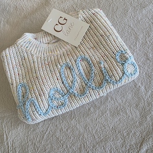 May include: A cream-colored knit sweater featuring a speckled multi-colored pattern and the word "hello" embroidered in light blue yarn across the front. A white rectangular brand tag with the text "CG lane" is attached to the neckline.