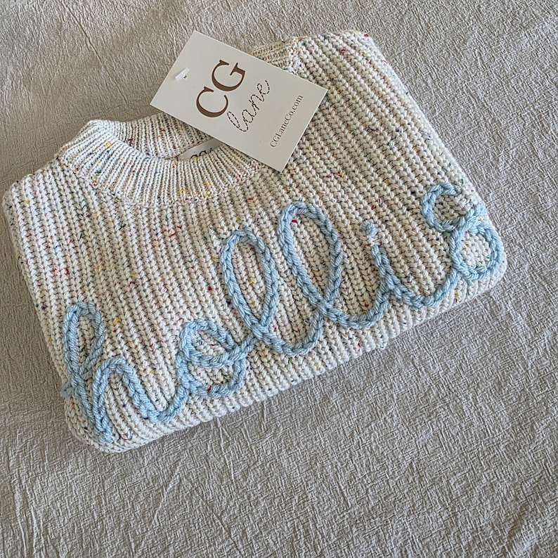 May include: A cream-colored knit sweater featuring a speckled multi-colored pattern and the word "hello" embroidered in light blue yarn across the front. A white rectangular brand tag with the text "CG lane" is attached to the neckline.