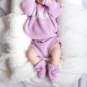 May include: A light purple knitted long-sleeve sweater with white cursive text on the chest, matching knitted shorts with a drawstring waist, and a pair of matching knitted booties with tie closures, all featuring a ribbed texture throughout.