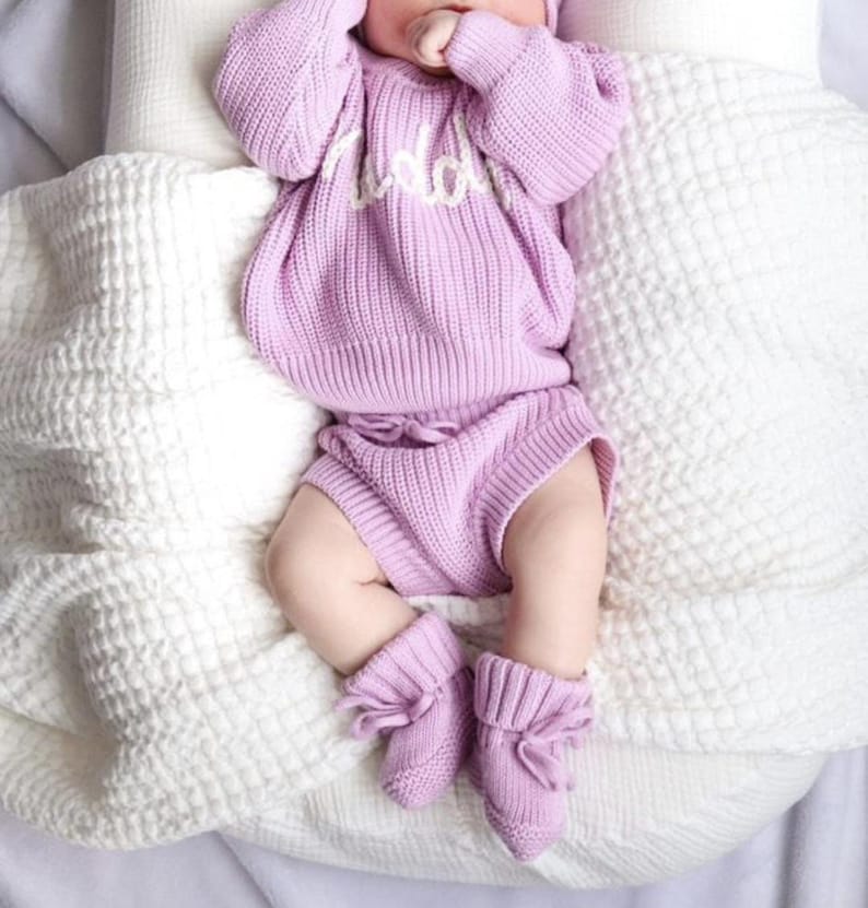 May include: A light purple knitted long-sleeve sweater with white cursive text on the chest, matching knitted shorts with a drawstring waist, and a pair of matching knitted booties with tie closures, all featuring a ribbed texture throughout.