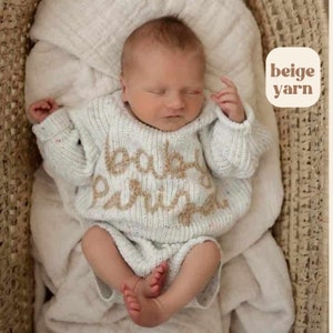 May include: A cream-colored knitted long-sleeve sweater and matching shorts set featuring the embroidered text "baby" and "pariza" in a tan script font on the chest. The knit fabric has subtle multicolored flecks throughout the material.