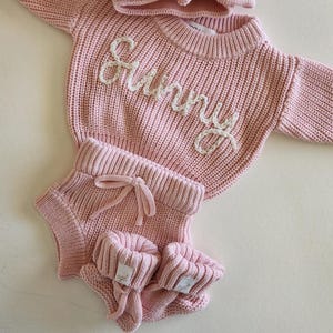 May include: A light pink knitted long-sleeve sweater with the word Sunny embroidered in white yarn across the chest, paired with matching knitted shorts featuring a drawstring waist and a pair of small knitted booties with white brand labels.
