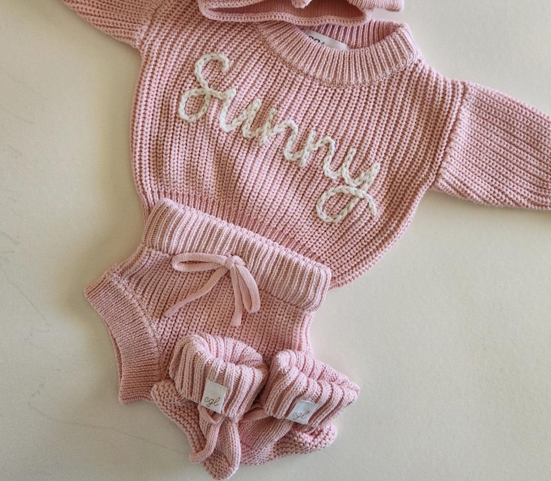 May include: A light pink knitted long-sleeve sweater with the word Sunny embroidered in white yarn across the chest, paired with matching knitted shorts featuring a drawstring waist and a pair of small knitted booties with white brand labels.