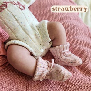 May include: A cream-colored knit romper with multicolored flecks and a pink embroidered strawberry design, paired with matching light pink knit booties featuring tie closures. The romper has a ribbed texture and white buttons at the leg opening.