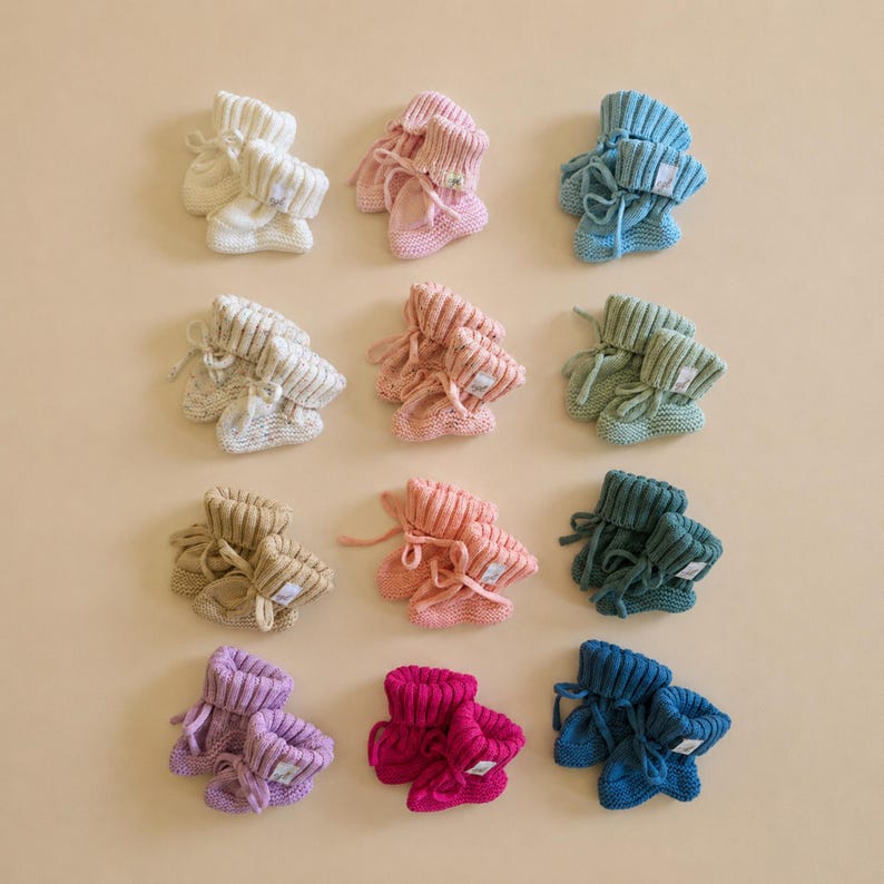 May include: Twelve pairs of knitted baby booties arranged in a grid. The booties feature a ribbed cuff and a tie closure, appearing in various solid colors including cream, pink, light blue, beige, sage green, dark teal, purple, and magenta.