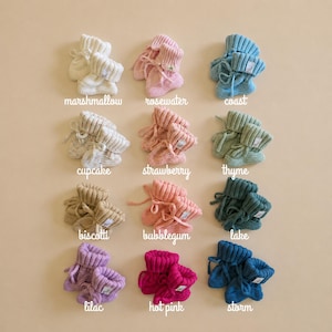 May include: A collection of twelve pairs of knitted baby booties arranged in a grid, each labeled with color names including marshmallow, rosewater, coast, cupcake, strawberry, thyme, biscotti, bubblegum, lake, lilac, hot pink, and storm.