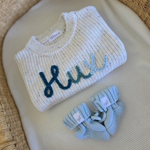 May include: A cream-colored knit sweater with multicolored flecks and the name Hux embroidered in a gradient of blue yarn across the front, paired with a matching pair of light blue knit baby booties featuring small white brand labels.
