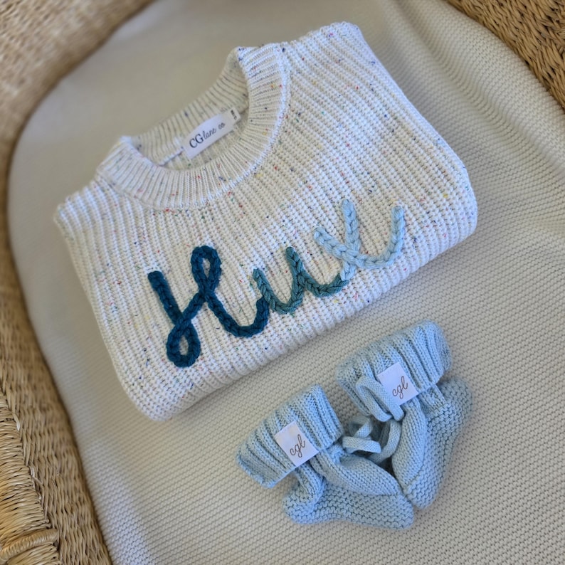 May include: A cream-colored knit sweater with multicolored flecks and the name Hux embroidered in a gradient of blue yarn across the front, paired with a matching pair of light blue knit baby booties featuring small white brand labels.