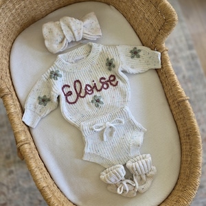 May include: A cream-colored knit sweater and matching bloomers set with a speckled texture, featuring the name Eloise embroidered in dark red script, small green floral accents, a matching knit headband, and a pair of knit booties with ties.