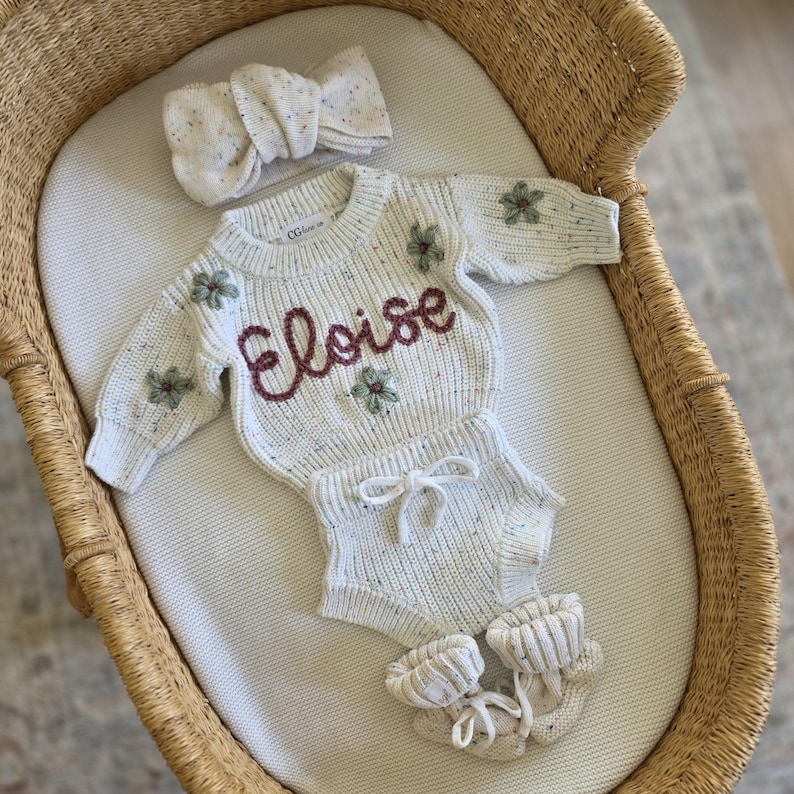 May include: A cream-colored knit sweater and matching bloomers set with a speckled texture, featuring the name Eloise embroidered in dark red script, small green floral accents, a matching knit headband, and a pair of knit booties with ties.