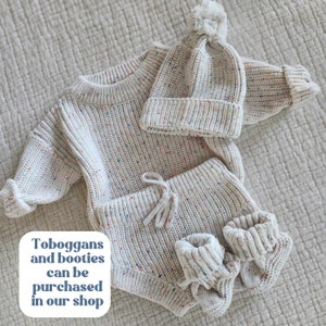 May include: A cream-colored knit sweater, matching shorts with a drawstring, a beanie with a pom-pom, and two booties, all featuring a multi-colored speckled pattern. The text on the image reads: Toboggans and booties can be purchased in our shop.