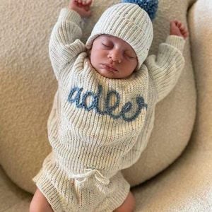 May include: A cream-colored knit sweater and matching beanie set featuring a speckled texture and the name Adler embroidered in blue cursive script across the chest. The sweater includes long sleeves and the bottoms feature a drawstring waist.
