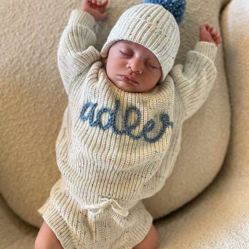 May include: A cream-colored knit sweater and matching beanie set featuring a speckled texture and the name Adler embroidered in blue cursive script across the chest. The sweater includes long sleeves and the bottoms feature a drawstring waist.