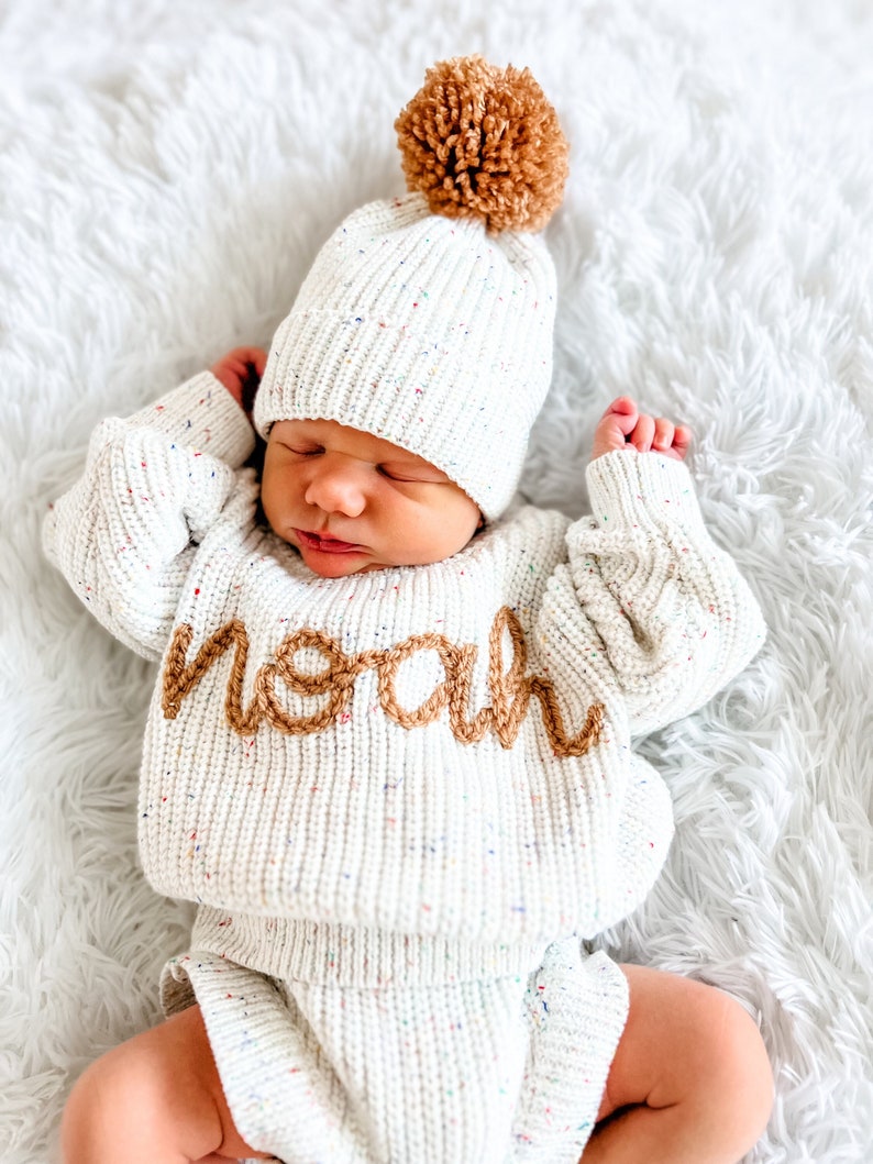 May include: A cream-colored knit sweater and matching beanie set with multicolored speckles. The sweater features the name "noah" embroidered in brown yarn across the chest. The beanie is topped with a large, textured brown yarn pom-pom.
