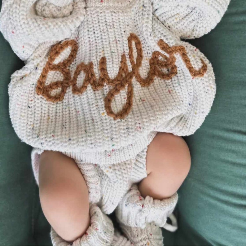 May include: A cream-colored knit sweater and matching knit bottoms with speckled multi-colored flecks. The sweater features the name Baylor embroidered in brown yarn across the chest in a cursive script. The set includes matching knit booties.
